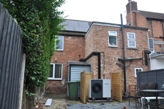 Back of a house with solar panels on the roof and an air source heat pump on the ground which is surrounded by wooden fence.